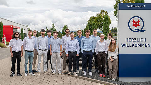 Eine Gruppe von dreizehn Personen, meist Männer, steht im Freien auf einem gepflasterten Weg neben einem großen Willkommensschild mit der Aufschrift "Herzlich Willkommen". Im Hintergrund sind Bäume und Gebäude unter einem bewölkten Himmel zu sehen.