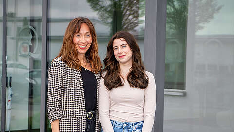 Zwei Frauen stehen dicht beieinander und lächeln in die Kamera vor einem modernen Gebäude mit großen Fenstern und roter Verkleidung.