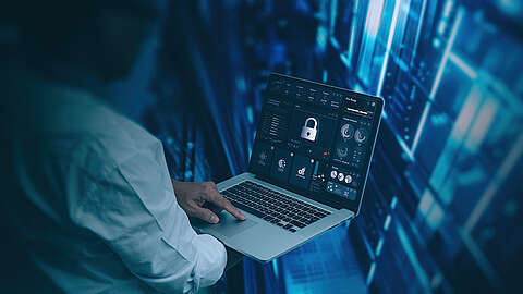 Eine Person, die einen Laptop benutzt, auf dem Symbole für Cybersicherheit und Datenschutz mit einem Vorhängeschloss-Symbol auf dem Bildschirm angezeigt werden, steht vor einem digitalen blauen Hintergrund, der Server oder Datennetze darstellt.