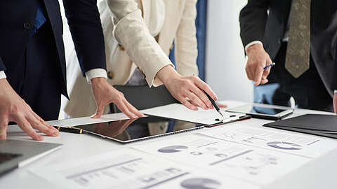 Drei Personen in Geschäftskleidung stehen um einen Tisch herum und sehen sich mit Tablets und einem Stift in der Hand Tabellen und Diagramme auf ausgedruckten Unterlagen an und besprechen diese. Der Fokus liegt auf ihren Händen und den Unterlagen.