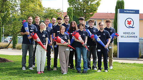 Eine Gruppe von Menschen in passenden marineblauen Hemden steht im Freien, lächelt und hält rote und blaue kegelförmige Geschenke in der Hand. Auf einem Schild neben ihnen steht "HERZLICH WILLKOMMEN" mit dem Q-FOX®-Logo. Im Hintergrund sind Bäume und ein Gebäude zu sehen.