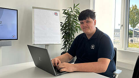 Ein junger Mann in einem dunklen Poloshirt sitzt an einem Schreibtisch und tippt auf einem Laptop. Er schaut in die Kamera und lächelt. In dem hellen Büroraum gibt es ein Flipchart, eine Pflanze und große Fenster.