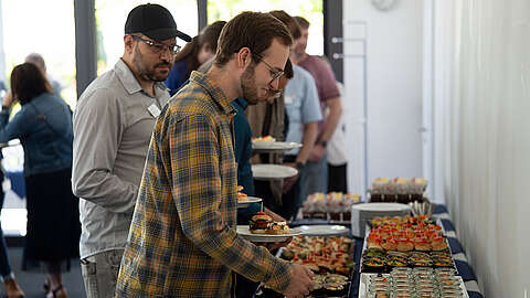 Mehrere Personen stehen in einer Schlange an einem Büffettisch und bedienen sich bei einer zwanglosen Veranstaltung im Haus. Der Tisch ist mit verschiedenen Gerichten und Desserts gefüllt, und die Leute halten Teller und Utensilien in der Hand.