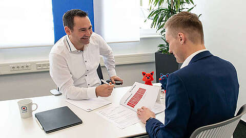 Zwei Männer in Geschäftskleidung sitzen sich an einem Schreibtisch gegenüber, lächeln und besprechen Dokumente. Ein Mann hält ein Papier mit Diagrammen in der Hand; auf dem Tisch stehen Tassen, Papierkram und ein Laptop. Zur Büroausstattung gehören Pflanzen und Plüschtiere.