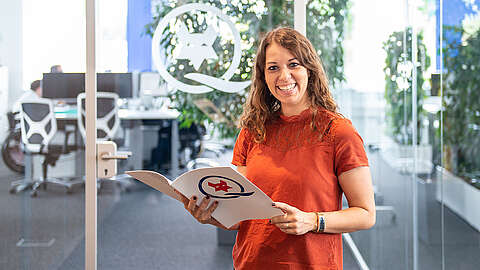 Lächelnde Frau vor einer Glaswand mit einer Q-FOX® Broschüre in der Hand