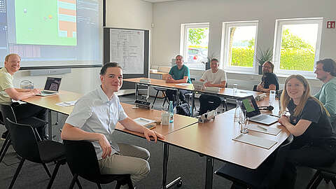 In einem hellen Besprechungsraum sitzen sieben Personen an Tischen, einige mit Laptops und Papieren. Auf einem großen Bildschirm wird ein Computerspiel gezeigt. Drei Fenster lassen Tageslicht herein, und im Hintergrund ist ein Whiteboard zu sehen.