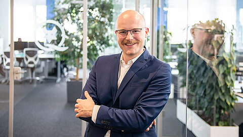 Ein lächelnder Mann mit Brille und Schnurrbart, gekleidet in einen blauen Anzug, steht mit verschränkten Armen in einem modernen Büro mit Glaswänden und Pflanzen. Sein Spiegelbild ist auf dem Glas neben ihm zu sehen.