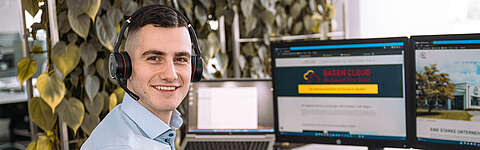 Q-FOX® Mitarbeiter sitzt an einem Schreibtisch mit PC-Monitoren und hat ein Headset auf
