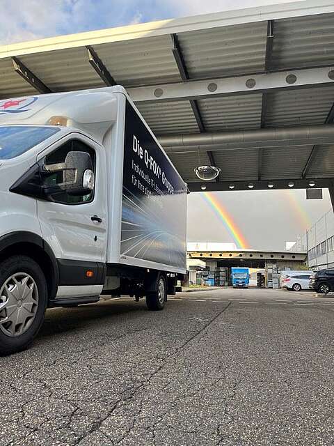 Ein weißer Kastenwagen ist unter einem Metalldach auf einem Industriegelände geparkt. Im Hintergrund spannt sich ein doppelter Regenbogen über den wolkenverhangenen Himmel, wobei Gebäude und ein weiterer blauer Lkw zu sehen sind.