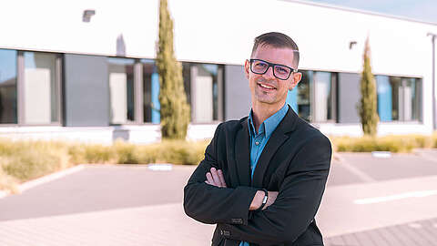 Für einen Quereinstieg in die IT ist man nicht zu alt Ein Mann mit Brille und schwarzem Anzug steht mit verschränkten Armen im Freien und lächelt in die Kamera. Im Hintergrund sind ein modernes Gebäude, Bäume und ein sonniger, klarer Himmel zu sehen.