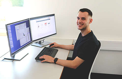 Karriereblogbeitrag - Yannik Merkel Junger Mann, der an einem Schreibtisch sitzt und in die Kamera lächelt. Er arbeitet an einem Computer mit zwei großen Monitoren.