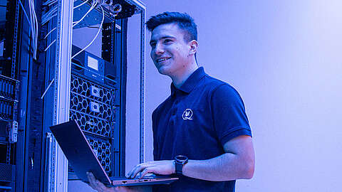 Ein junger Mann in einem marineblauen Polohemd steht in einem Serverraum und arbeitet lächelnd an einem Laptop. Hinter ihm sind Serverschränke und Kabel zu sehen, die alle von blauem Licht angestrahlt werden.