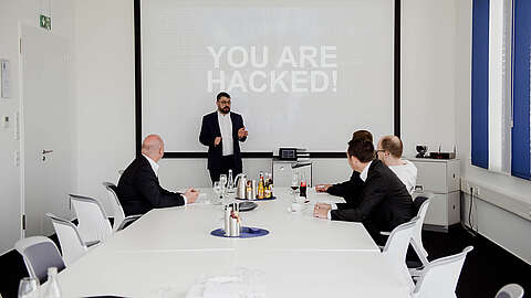 Ein Mann im Anzug steht vor einer Projektionsfläche mit der Aufschrift "YOU ARE HACKED!", während vier Personen in Geschäftskleidung an einem Konferenztisch sitzen und aufmerksam zuhören und zusehen.