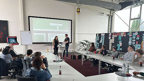 Eine Gruppe von Personen sitzt um einen Konferenztisch und beobachtet zwei Moderatoren, die vorne an einem großen Bildschirm stehen, auf dem eine Karte zu sehen ist. Notizen und Diagramme bedecken Tafeln an der Wand. Auf den Tischen stehen Flaschen mit Wasser und Limonade.