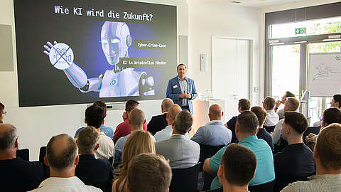 Ein Mann hält vor einem Publikum in einem Konferenzraum einen Vortrag. Auf einer Leinwand hinter ihm sind ein Roboter und ein deutscher Text über KI und Cyberkriminalität zu sehen. Die Teilnehmer verfolgen den Vortrag aufmerksam.