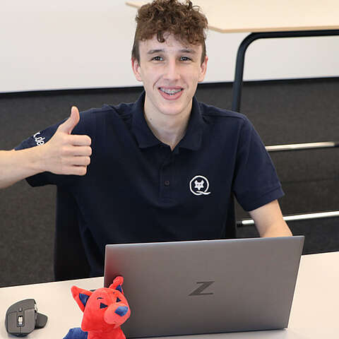Auszubildender sitzt mit seinem Notebook am Schreitisch und zeigt glücklich mit dem Daumen nach oben. Vor dem Notebook befindet sich ein roter Q-FOX® 