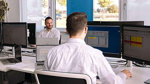 Drei Männer in weißen Hemden arbeiten an höhenverstellbaren Schreibtischen mit mehreren Computermonitoren in einem modernen Büro mit großen Fenstern und Zimmerpflanzen. Ein Mann blickt in die Kamera, während die anderen auf ihre Bildschirme konzentriert sind.