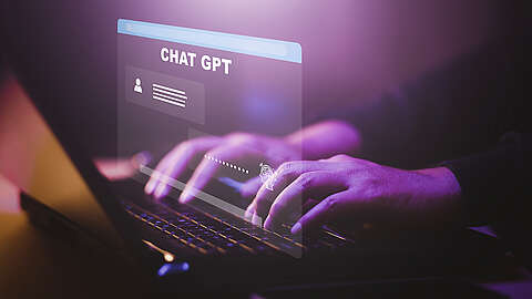 Hände, die auf einer Laptop-Tastatur tippen, mit einem leuchtenden virtuellen Bildschirm, auf dem "CHAT GPT" und Anmeldefelder angezeigt werden, was eine Interaktion mit einem KI-Chatbot nahelegt. Die Szene ist mit lila Licht beleuchtet.