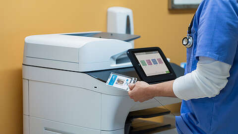 Eine Person in blauem Kittel mit einem Stethoskop hält einen Ausweis in der Nähe eines großen Bürodruckers oder -kopierers und benutzt ihn möglicherweise, um auf das Gerät zuzugreifen oder es in einer professionellen Umgebung zu bedienen.