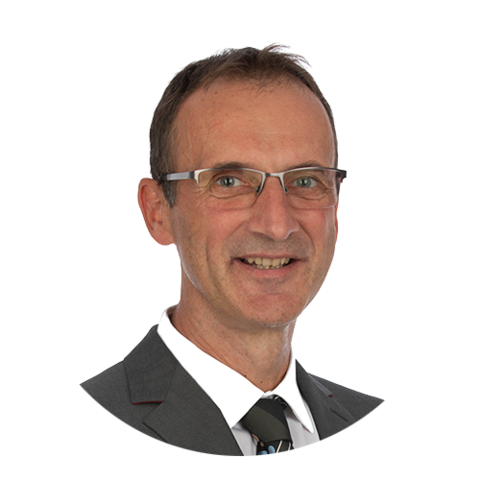 Lächelnder Mann mit Brille, dunklem Anzug, weißem Hemd und gemusterter Krawatte, vor einem schlichten weißen Hintergrund.