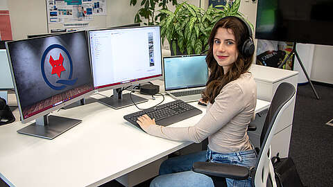 Eine Frau mit langen braunen Haaren, die Kopfhörer und ein beigefarbenes Oberteil trägt, sitzt an einem Schreibtisch mit zwei Monitoren und einem Laptop, tippt auf einer Tastatur und blickt in die Kamera in einer modernen Büroumgebung.