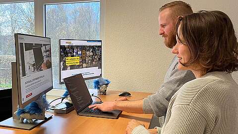 Den richtigen Partner für individuelle Softwareentwicklung finden Zwei Personen arbeiten an einem Schreibtisch mit zwei Monitoren und einem Laptop. Einer tippt, während der andere zusieht. Auf dem Schreibtisch liegen kleine blaue und graue Stofftiere. Vor dem Fenster sind Bäume zu sehen.