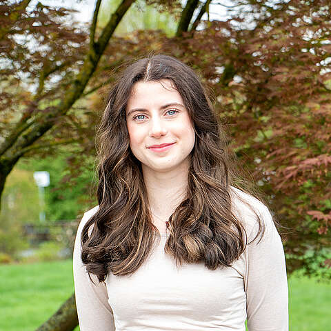 Eine junge Frau mit langen, gewellten braunen Haaren und einem hellbeigen Oberteil steht im Freien vor einem Baum mit rötlichen Blättern und lächelt sanft in die Kamera. Im Hintergrund sind grünes Gras und weitere Bäume zu sehen.