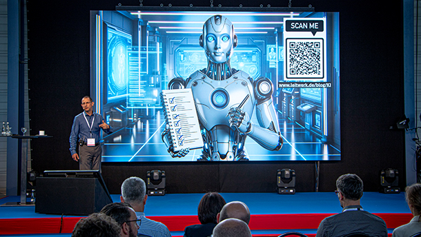 Ein Redner hält einen Vortrag vor einem Publikum, während hinter ihm auf einem großen Bildschirm ein humanoider Roboter zu sehen ist, der ein Dokument hält. Auf dem Bildschirm sind auch ein QR-Code und futuristische blaue Bilder zu sehen.