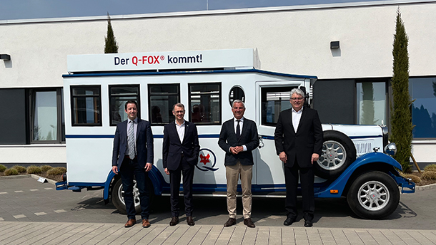 Vier Männer in Anzügen stehen vor einem weiß-blauen Oldtimer-Fahrzeug, auf dessen Dach ein Schild mit der Aufschrift "Der Q-FOX® kommt!" angebracht ist. Im Hintergrund sind ein modernes Gebäude und ein klarer Himmel zu sehen.