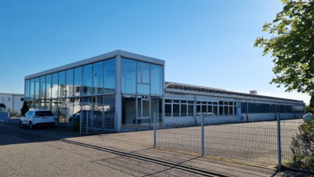 Ein modernes, verglastes Geschäftsgebäude mit großen Fenstern, einem Flachdach und einem eingezäunten Parkplatz. Ein weißes Auto ist in der Nähe des Eingangs geparkt. Der Himmel ist klar und blau.