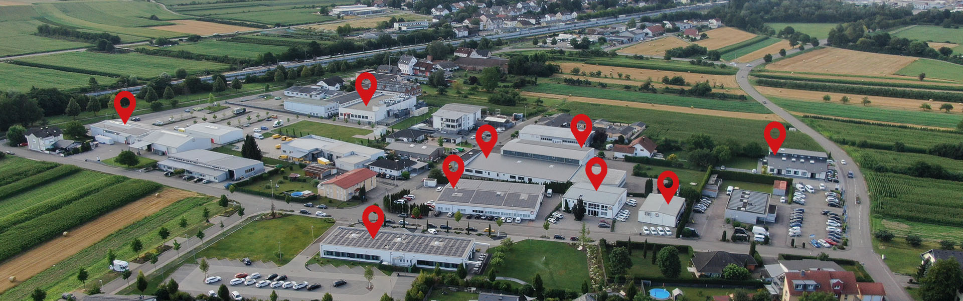 Luftaufnahme eines kleinen Industriegebiets mit mehreren Gebäuden und roten Stecknadeln über mehreren Gebäuden, umgeben von Feldern und einem entfernten Wohngebiet.
