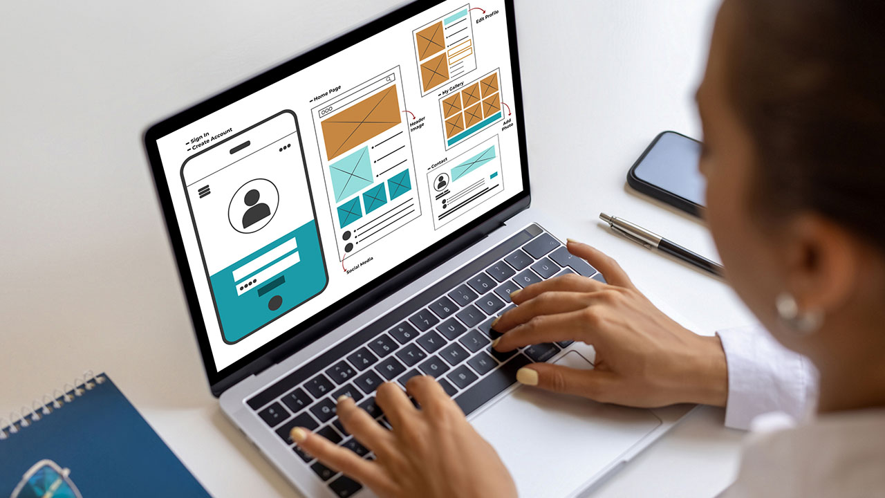 Eine Person arbeitet an einem Laptop und zeigt Wireframes und Benutzeroberflächendesigns für eine Website oder App an. Ein Notizbuch, ein Stift und ein Smartphone liegen in der Nähe auf dem weißen Schreibtisch.