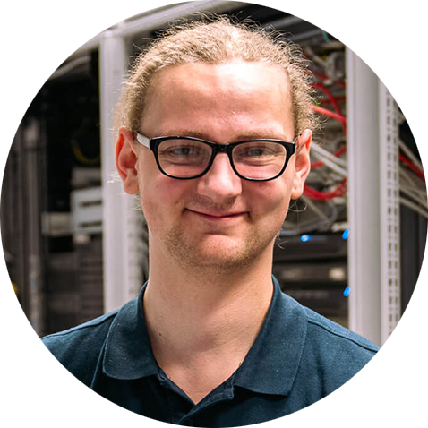 Eine Person mit heller Haut, Brille und zurückgebundenem, langem, hellem Haar, lächelnd und in einem dunklen Hemd mit Kragen steht vor Computerservern, im Hintergrund sind Kabel und Geräte sichtbar.