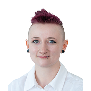 Eine Person mit heller Haut, kurzen, dunkelrosa Haaren, die zu einem Irokesenschnitt frisiert sind, rasierten Seiten, blauen Augen, Piercings im Gesicht und schwarzen Ohrringen, die ein weißes Hemd mit Kragen trägt, lächelt vor einem schlichten, hellen Hintergrund in die Kamera.