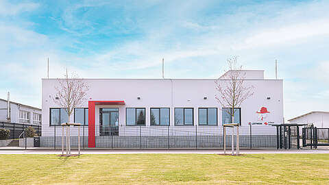 Hinter einem Zaun steht ein modernes weißes Gebäude mit großen Fenstern und einem roten Akzent am Eingang. Davor stehen zwei junge Bäume auf einer grünen Wiese unter einem teilweise bewölkten Himmel.