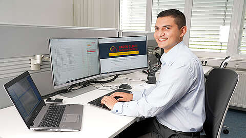 Junger Mitarbeiter sitzt an einem PC mit mehreren Bildschirmen und tippt in die Tastatur